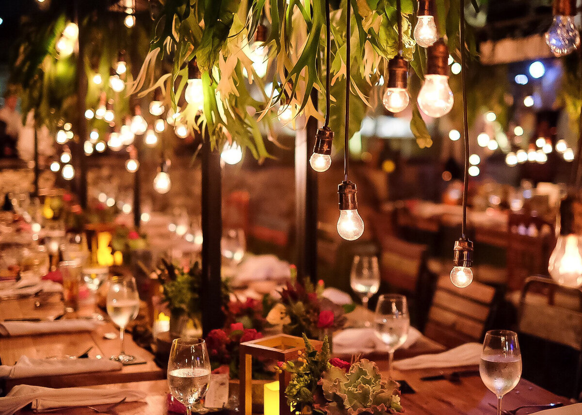 A dinner table set up for a luxury party with hanging lights and greenery outdoors.
