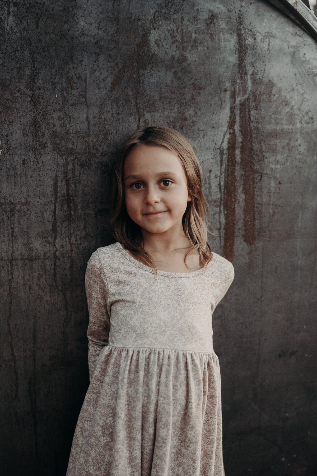 A close-up image of a little girl, wearing a long sleeve dress with shorter brown hair, and a mischievous smile on her face as she poses on her family's farm.