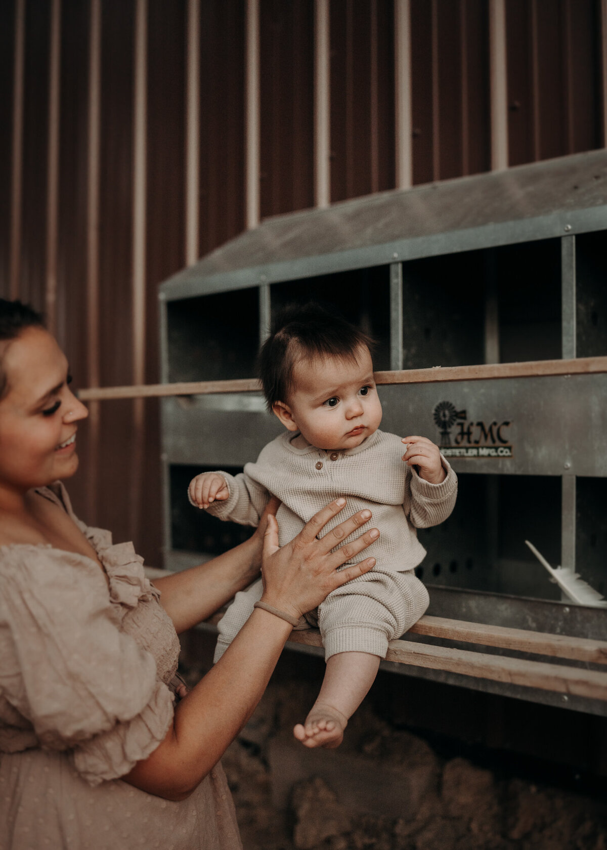 a mother smiles at her young baby as he sits on the ledge of their family's chickens nesting box in the fall at sunset.