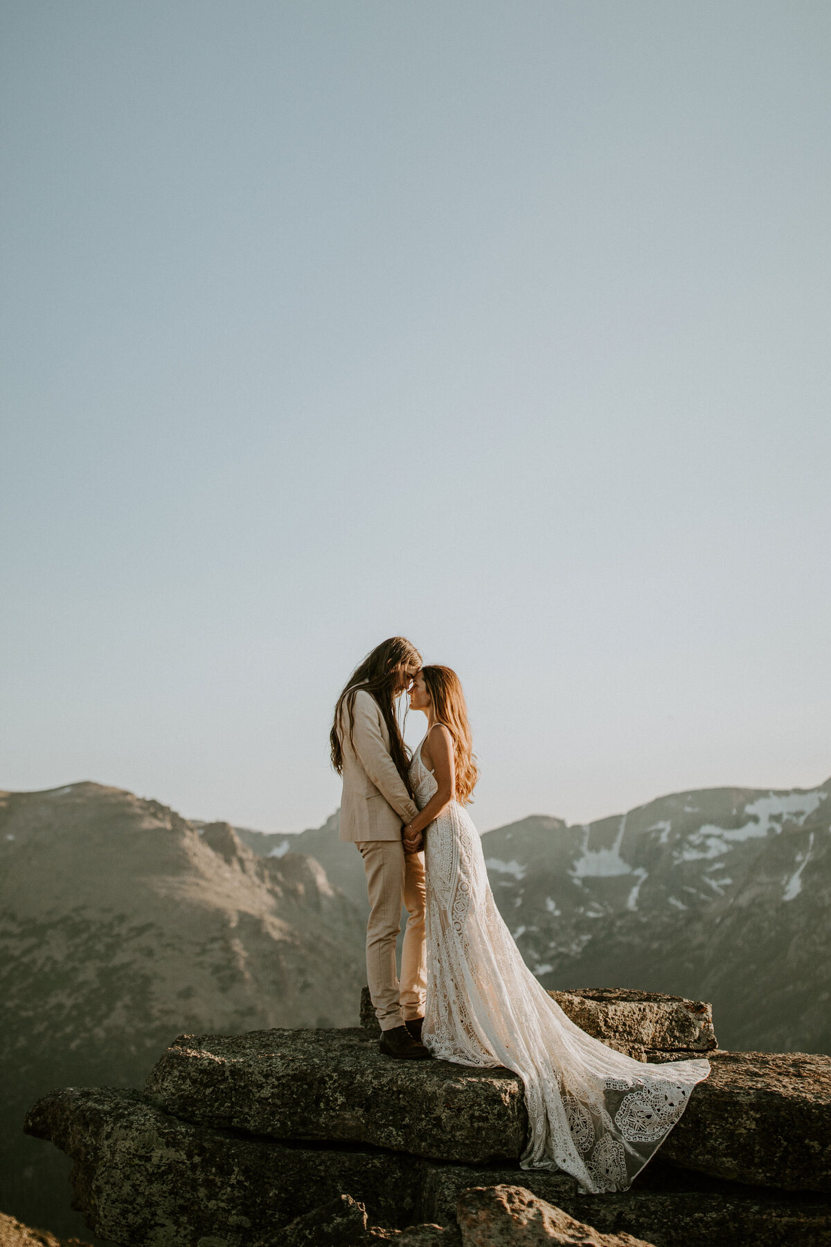 Bride and groom wearing an ivory suit and white wedding gown holding each other atop a stone in the mountains.