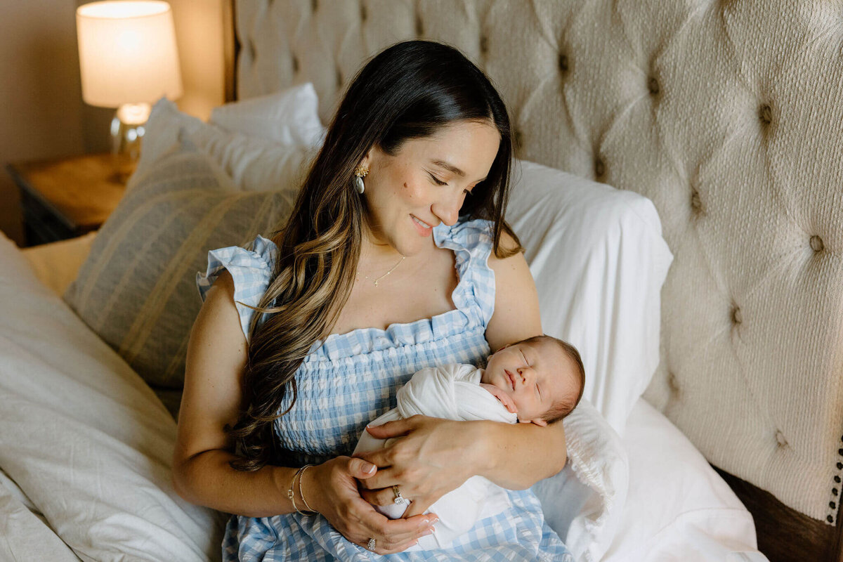 mother in blue and white plaid dress holding newborn swaddled in cream blanket while sitting on edge of bed