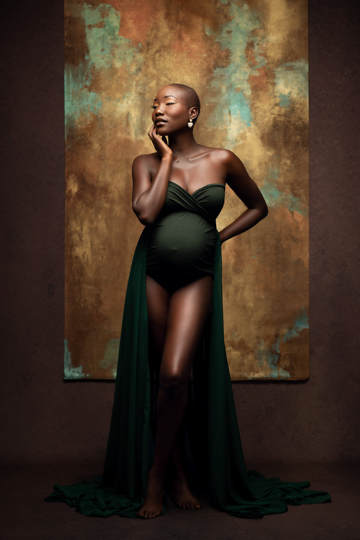 Stunning black, pregnant woman wearing a green bodysuit with green chiffon panels flowing on either side of her and a fine art backdrop behind.