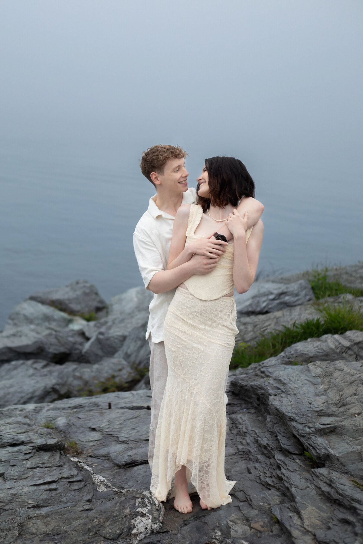 Engagement Photos Rhode Island | Kelsey Sheehan Photography Timeless Rhode Island Weddings | A couple embraces on rocky terrain by calm water. The man in a white shirt hugs the woman in a cream dress, both smiling. The mood is serene and intimate.