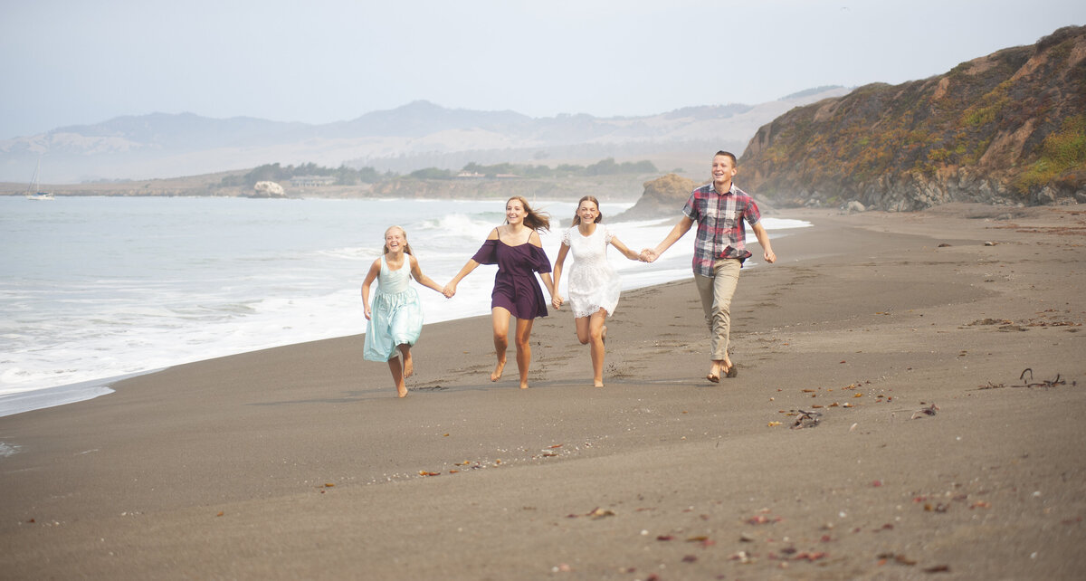 4 barefoot Kids running toward camera view and smiling while holding hands on the seashore