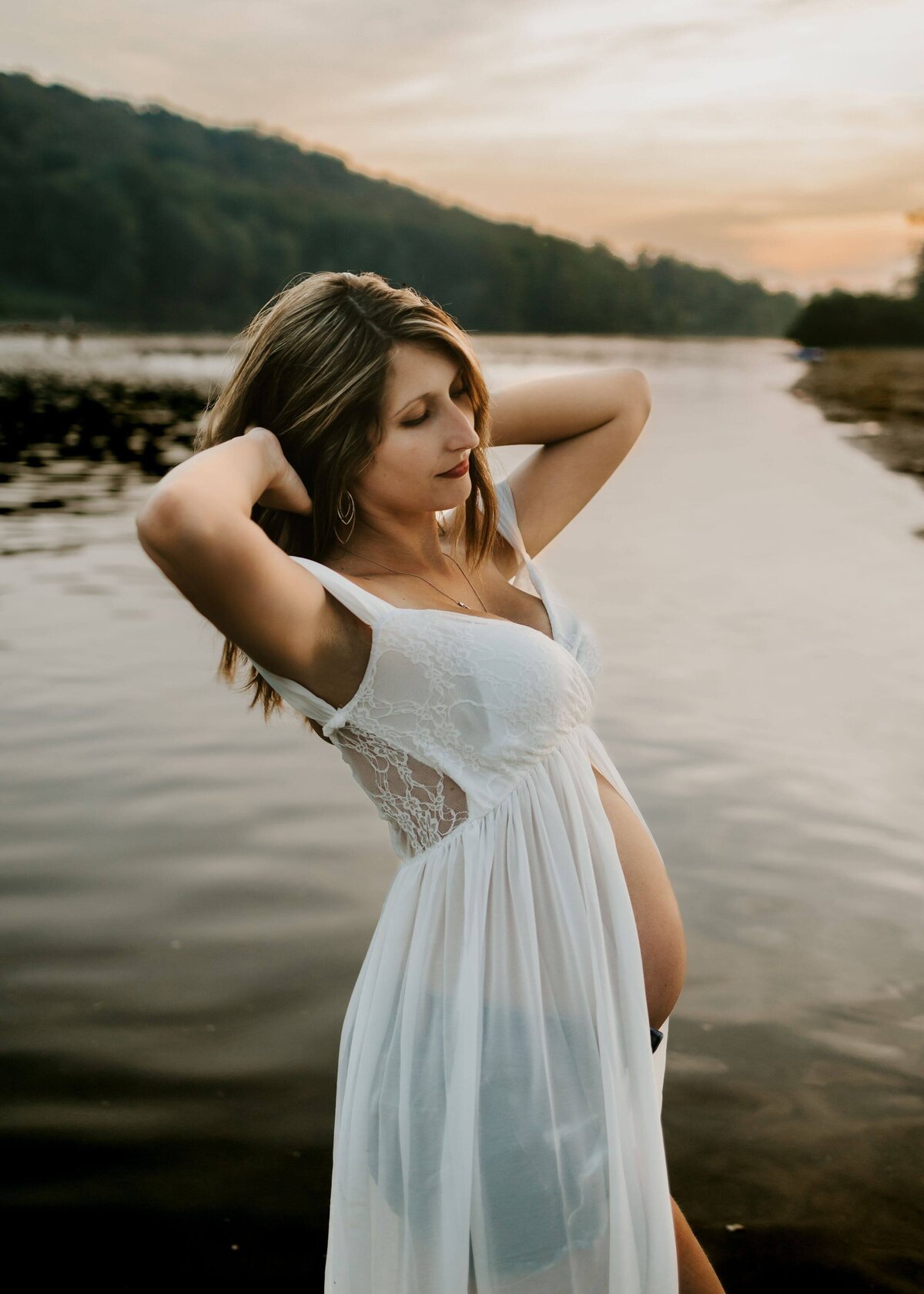 A pregnant woman in a white dress posing by the water captured by a Pittsburgh maternity photographer.