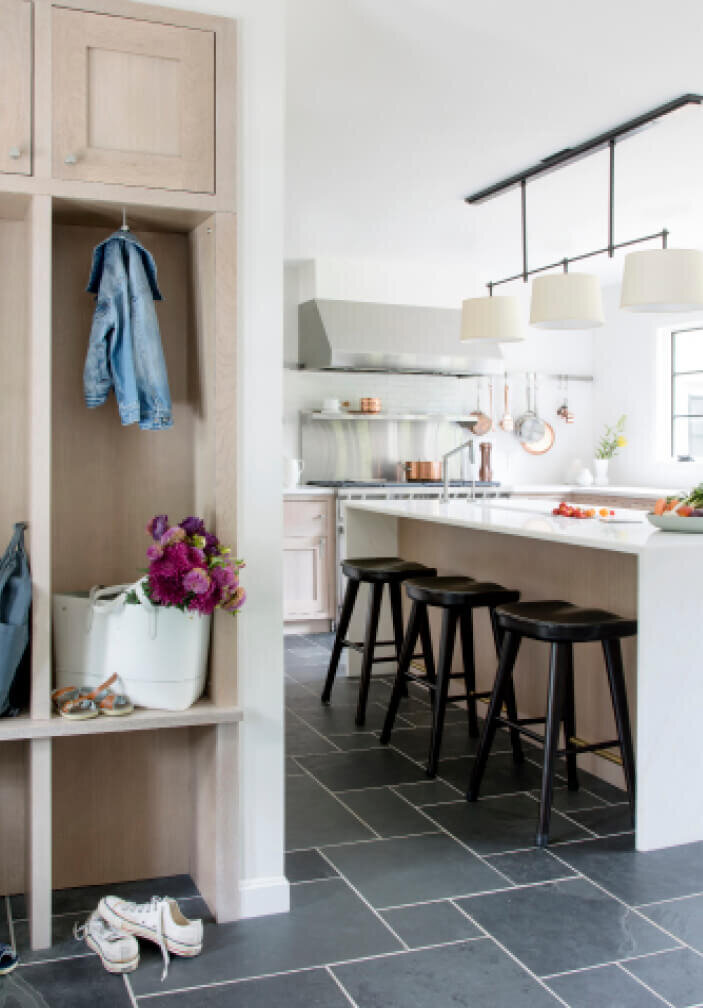 Kitchen and bath designers in Westwood create a renovation with natural oak cabinets, slate flooring, black tractor stools, and custom mudroom cubbies.