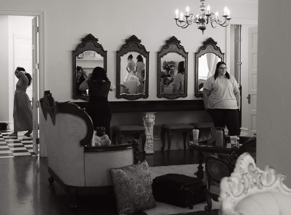 a documentary wedding photograph of the bridal suite during getting ready, in one of the mirrors, you can see the reflection of the bride getting her dress zipped up by her mom as a friend watches from one corner, the mother of the groom fixes her hair in another mirror and another friend walks around the bathroom getting jewelry on. by documentary wedding photographer - Primrose Wedding & Adventure Photography