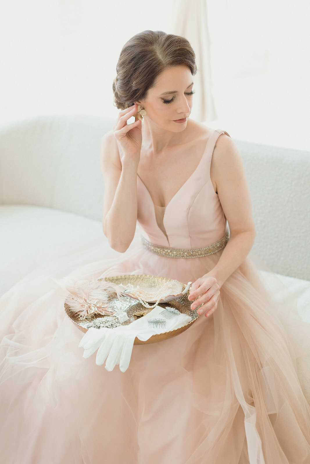 light skinned woman in pink ballgown is seated, with a tray of dressing items and jewelry, and holding one of the earrings to her ear..