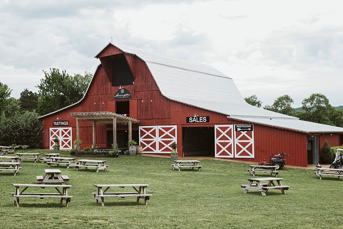 Large red barn with a hay loft and large white doors in a field with picnic benches.