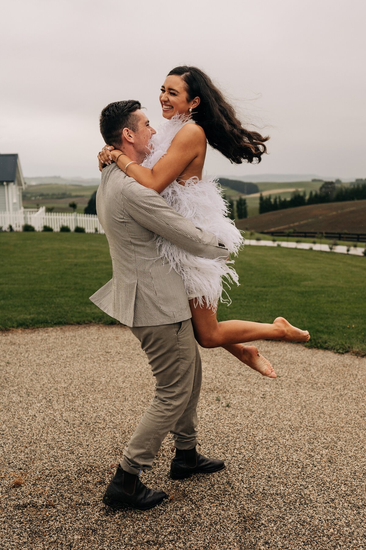 groom in chinos and boots and striped blazer picks up bride in feathered mini reception party dress and they laugh while spinning