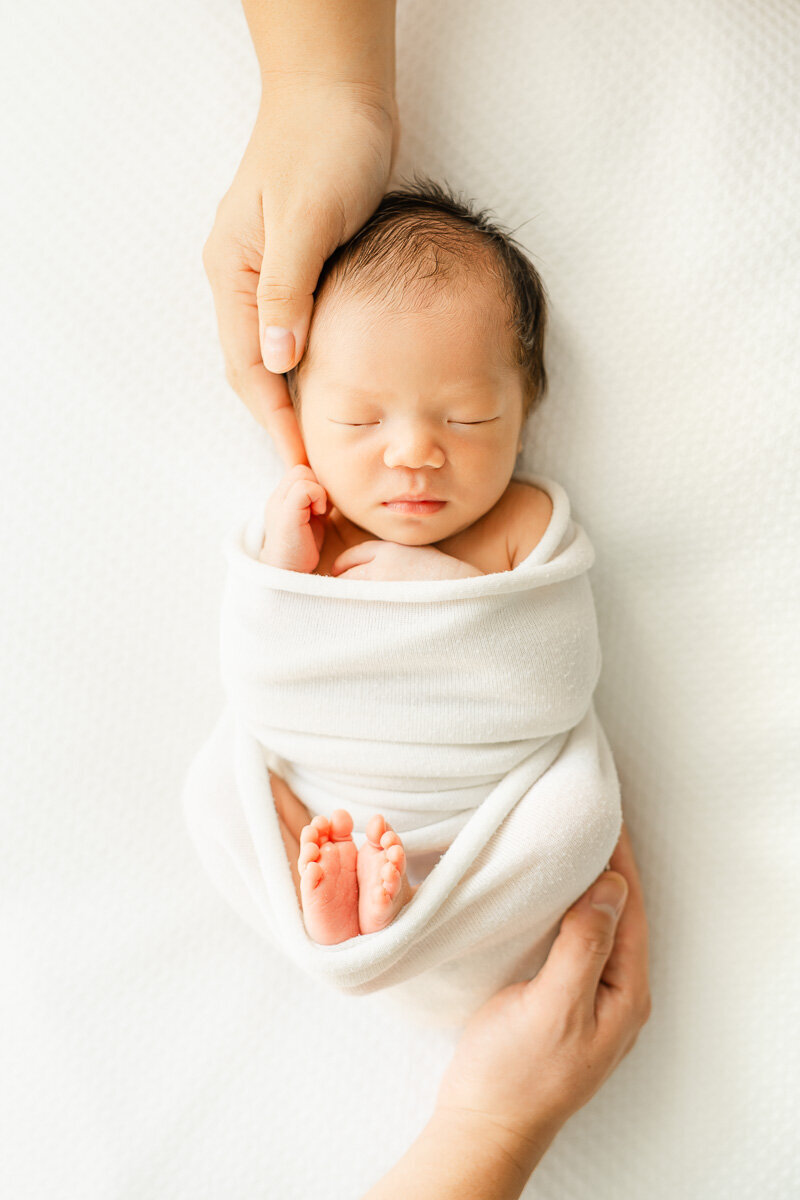 an infant child lays on a white bean bag while wrapped in a white blanket. One adult hand hold her head while another adult hand holds her feet in this shot captured by a newborn photographer in Austin, TX.