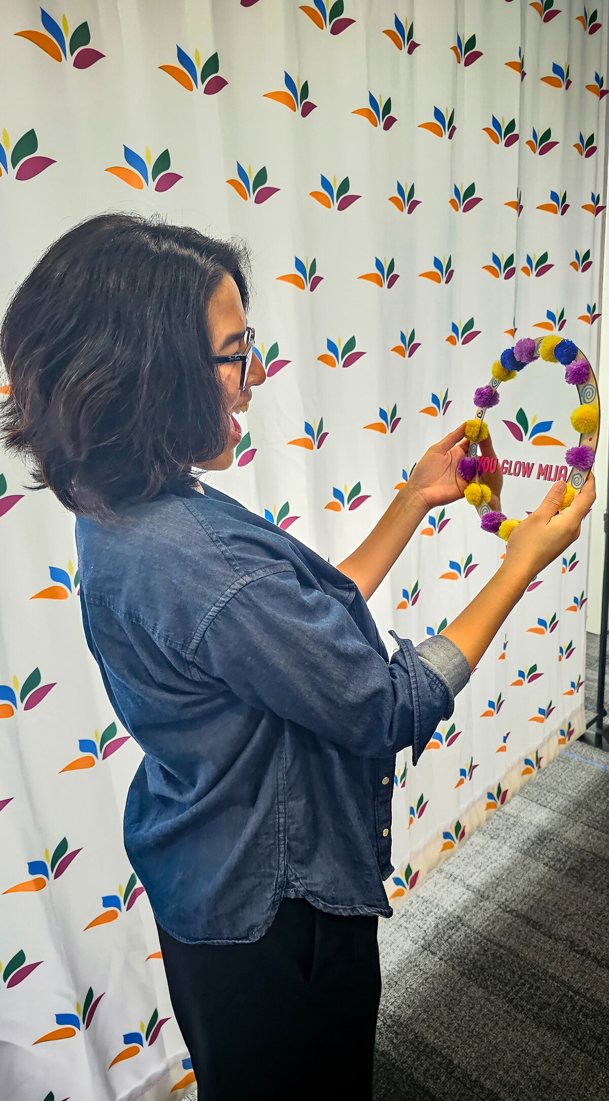 Woman proudly showcases her creation against a textured, multicolored backdrop. Behold a round mirror adorned with an array of materials, reflecting the essence of vibrant artistic expression.