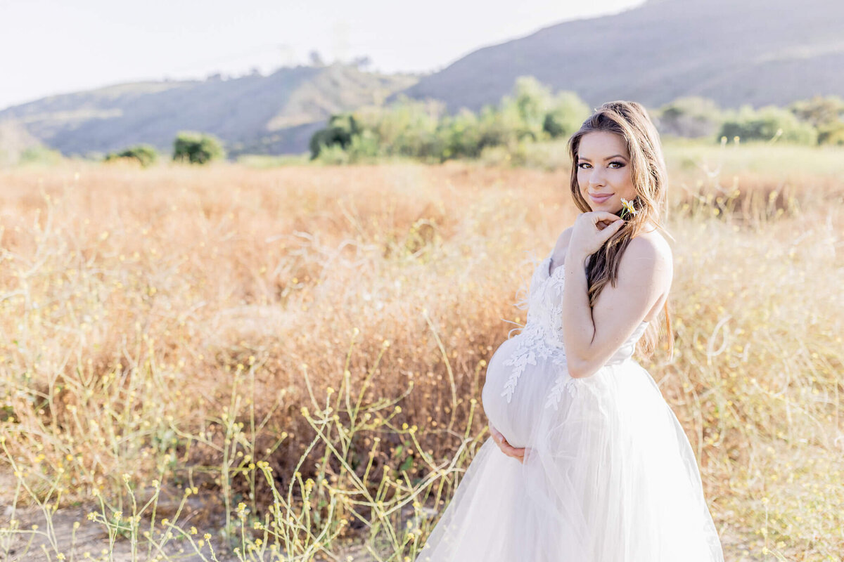 A pregnant woman in a white dress stands in a sunlit field, holding her belly with one hand and a flower in the other. Mountains are visible in the background.
