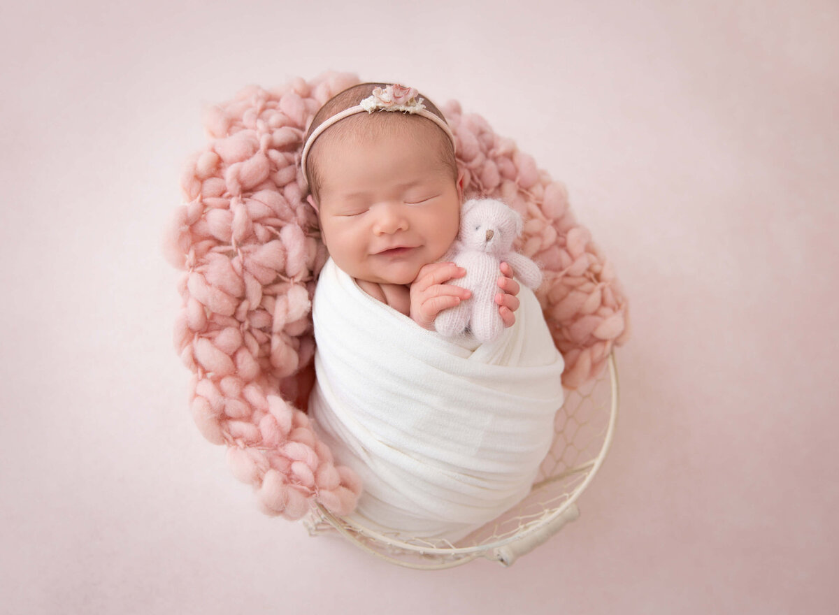 Newborn baby girl wrapped in soft white fabric, gently holding a blush teddy bear in her tiny hands, beautifully captured by a Brooklyn newborn photographer, showcasing the pure innocence of her early days.