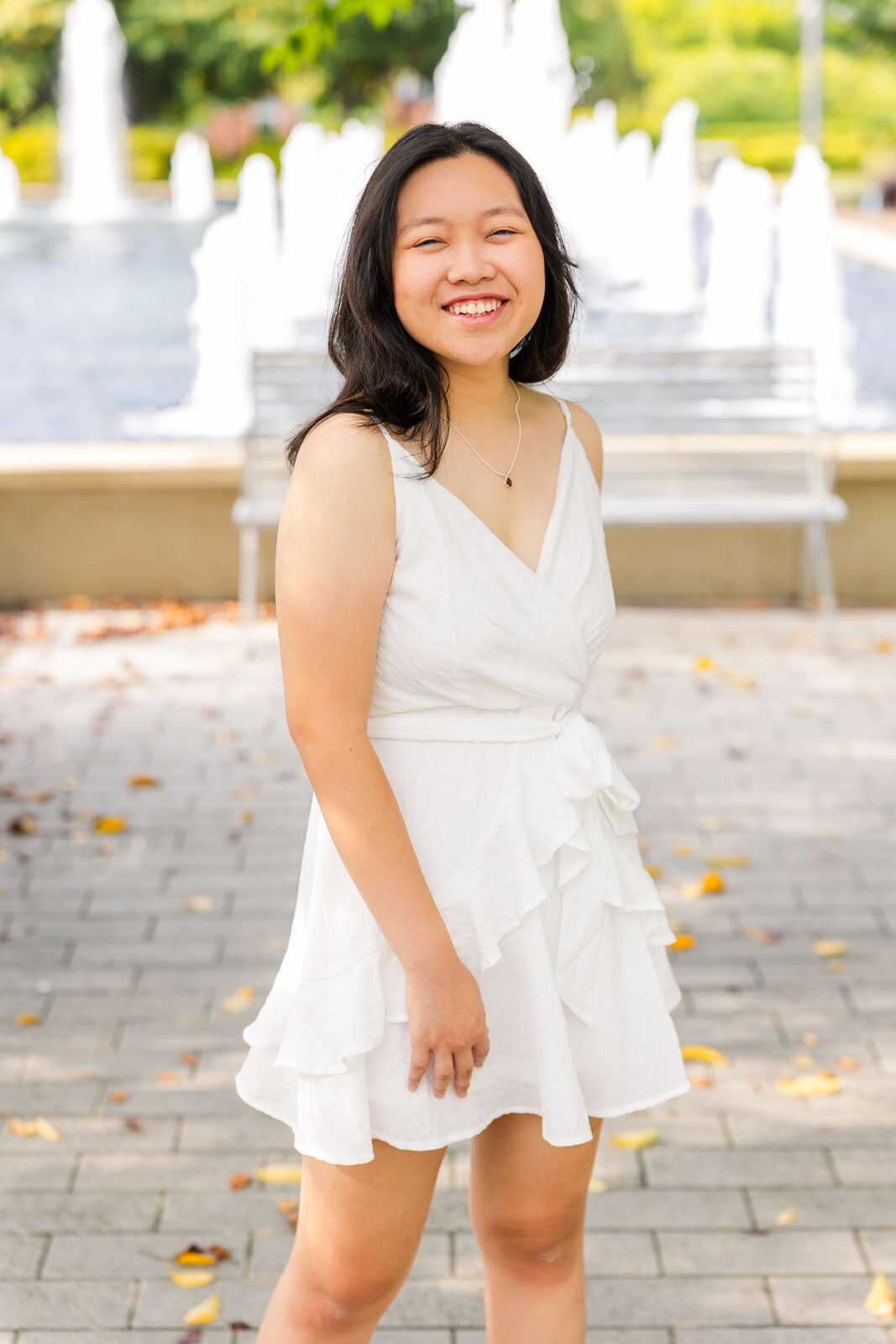 senior girl wearing a white short dress, laughing at the camera and twirling in front of a fountain in a urban environment during photoshoot