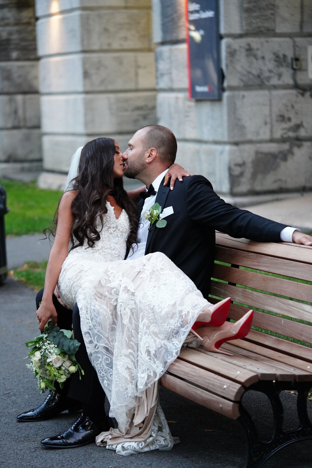 The bride sits on the groom's lap on a park bench, sharing a sweet kiss. Surrounded by a serene park setting, this intimate moment highlights their affection and connection amidst a relaxed, natural backdrop.