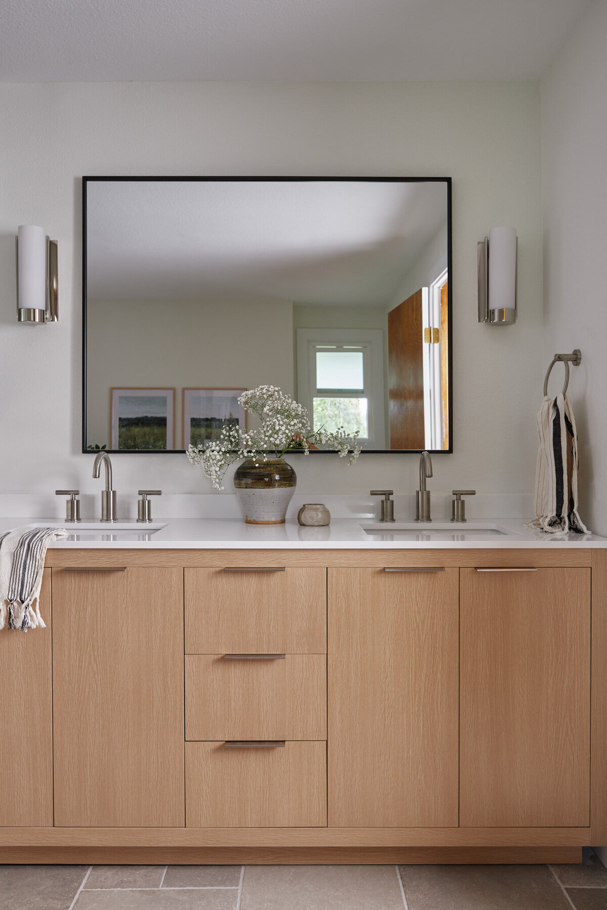 Bathroom with white oak vanity with silver cabinet pulls and white countertops with silver faucets. Large mirror with thin black frame and silver and white sconces on the sides. White and black striped towel draped over the sink. Antique vase with brown and white glaze and babys breath flowers in it. Cream colored jar on the vanity counter next to the flowers.