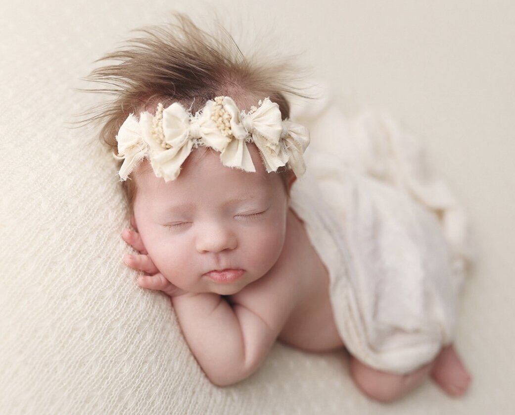 Baby girl posed on stomach and facing camera asleep with a light yellow blanket covering her back and wearing a yellow bow headband.