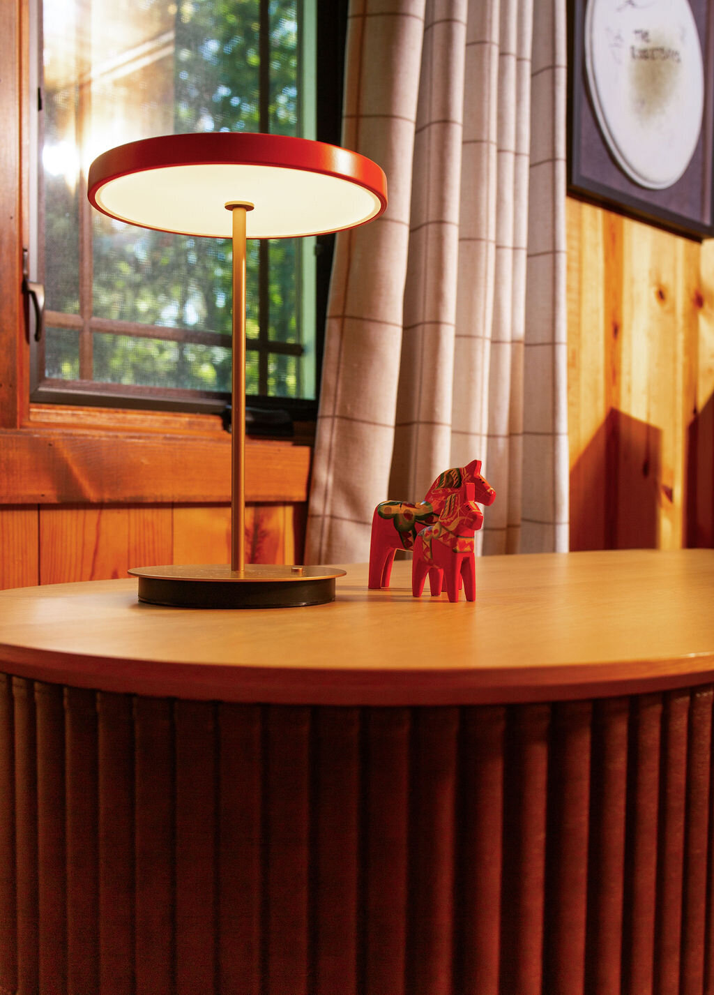 A close-up of a wooden tabletop with a distinctive ribbed edge. A modern red and gold table lamp with a circular shade illuminates the surface. A small, red wooden Dala horse figurine stands next to the lamp. A window with a view of greenery and a striped curtain is visible in the background. A framed circular object hangs on the wood-paneled wall.