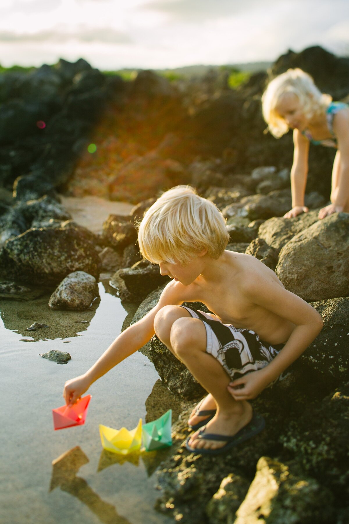 siblings play with paper boats in tidepools at makapuu beach at sunset