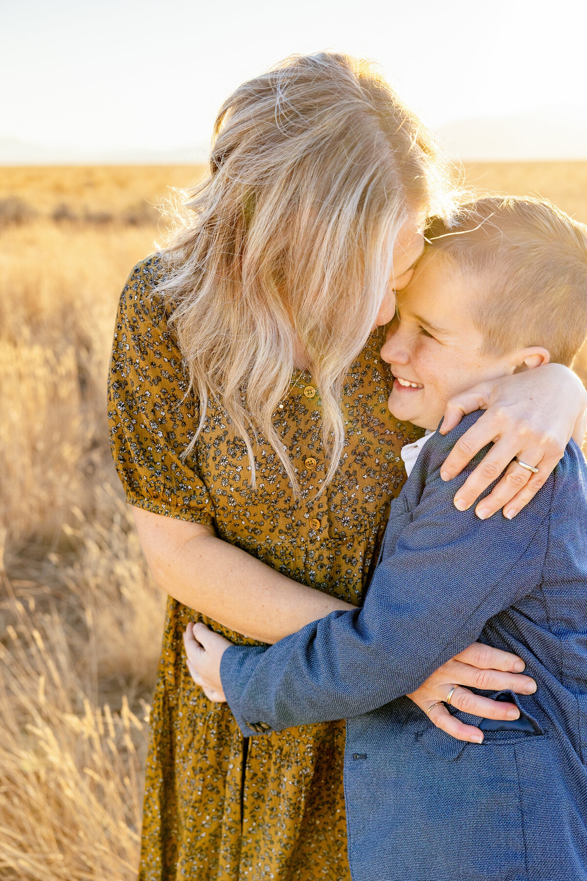 A mother embraces her young son, both smiling warmly as the golden sunlight illuminates their faces in an open field.
