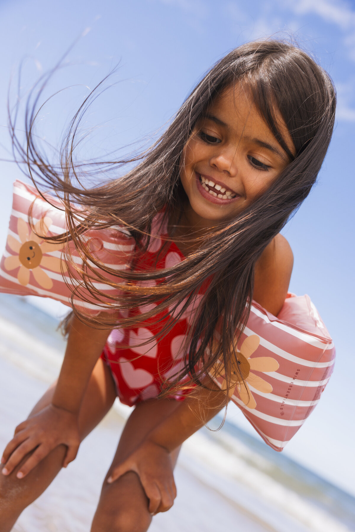 Smiling girl at the beach wearing a red heart-print swimsuit and pink striped Swim Essentials floaties with cheerful flower faces, with her hair blowing in the wind.