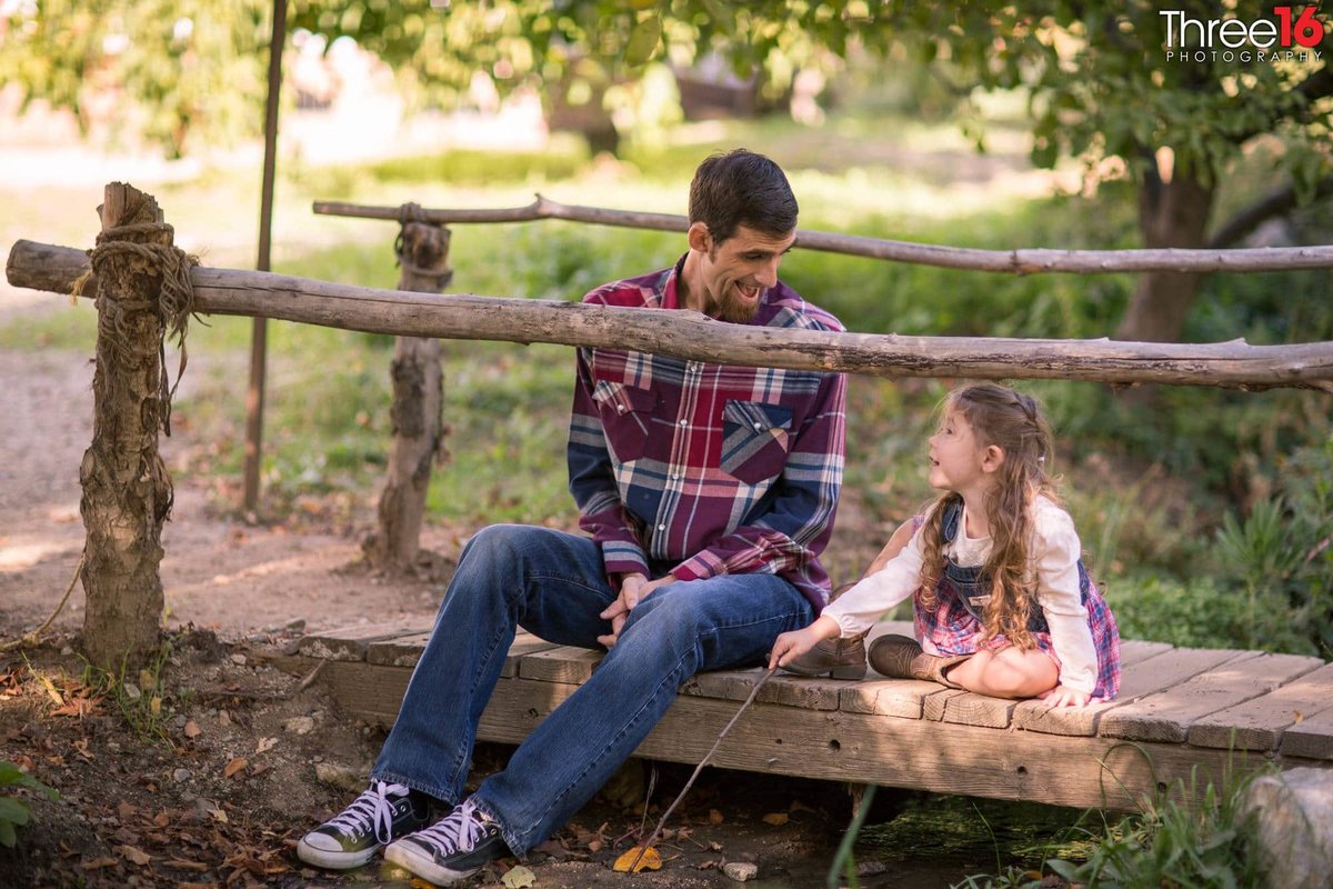 Father and daughter look at each other while sitting on a bridge over a small creek
