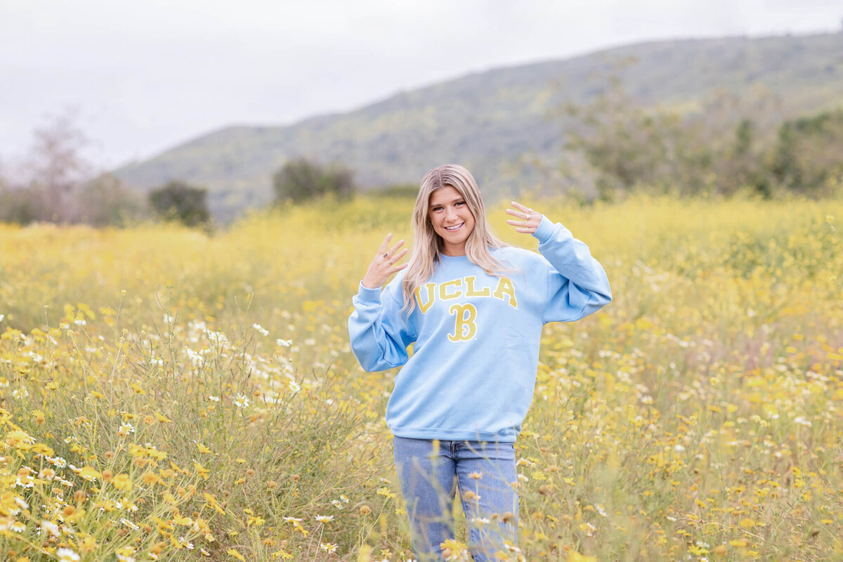 A person stands in a field of yellow wildflowers, smiling and showing a playful gesture with their hands. They are wearing a blue UCLA sweatshirt. Hills are visible in the background under a cloudy sky.