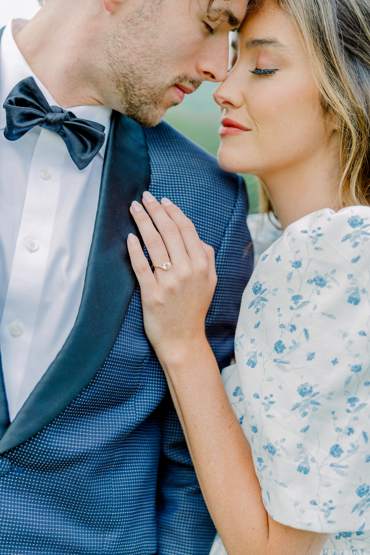 Ivy Rose Barn | A close up of a romantic bride and groom nuzzling close to one another. The groom wears a navy tuxedo, while the bride, in a vintage-inspired floral gown with puff sleeves, lays her hand on his chest. Captured by Virginia Wedding Photographer, Caitlin Wilcox.