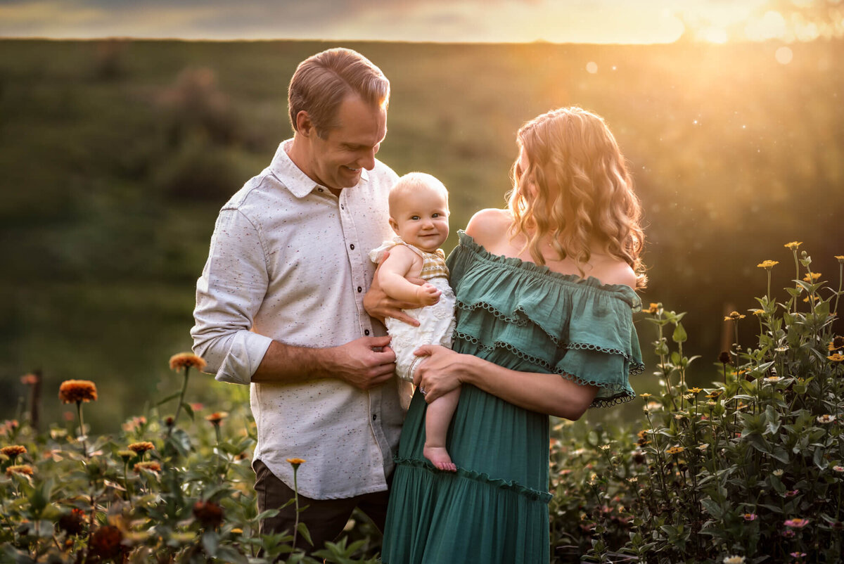 A mom in a green dress holds her baby girl in a field a flowers with dad cuddling too