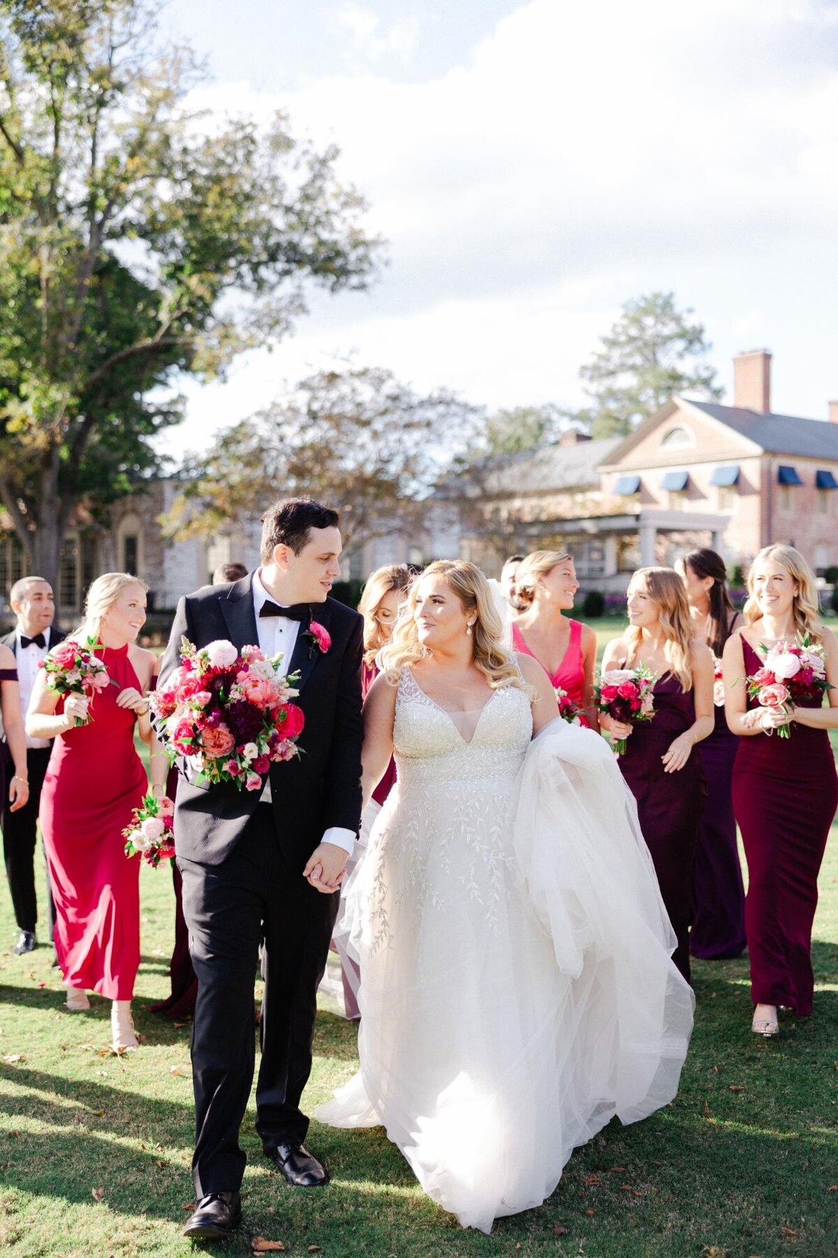 A bride and groom walking hand in hand, accompanied by bridesmaids in pink and red dresses.