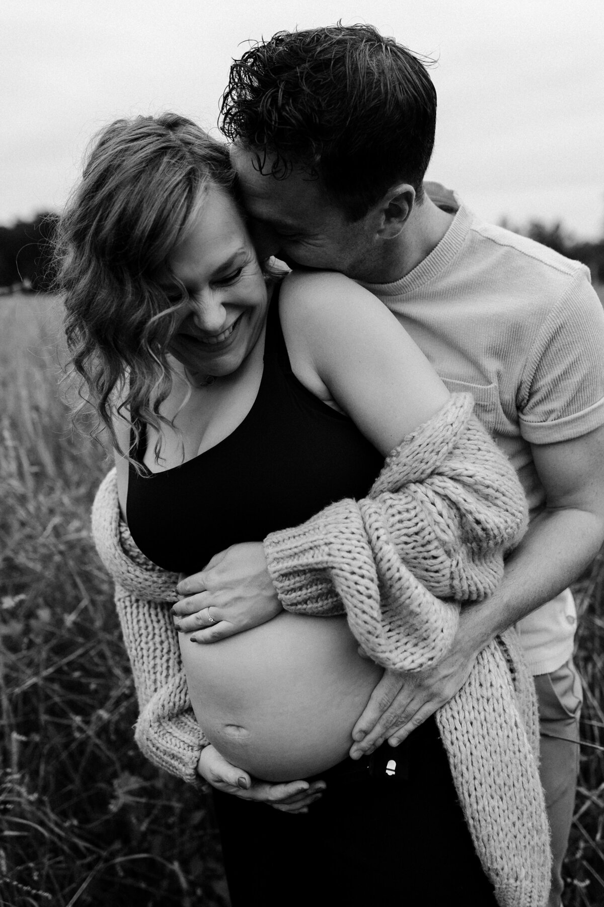 Foto door Jamie de Leeuw zwangerschapsshoot fotograaf man staat achter vrouw en fluistert iets in oor vrouw zwarte top blote buik en vest zwartwit