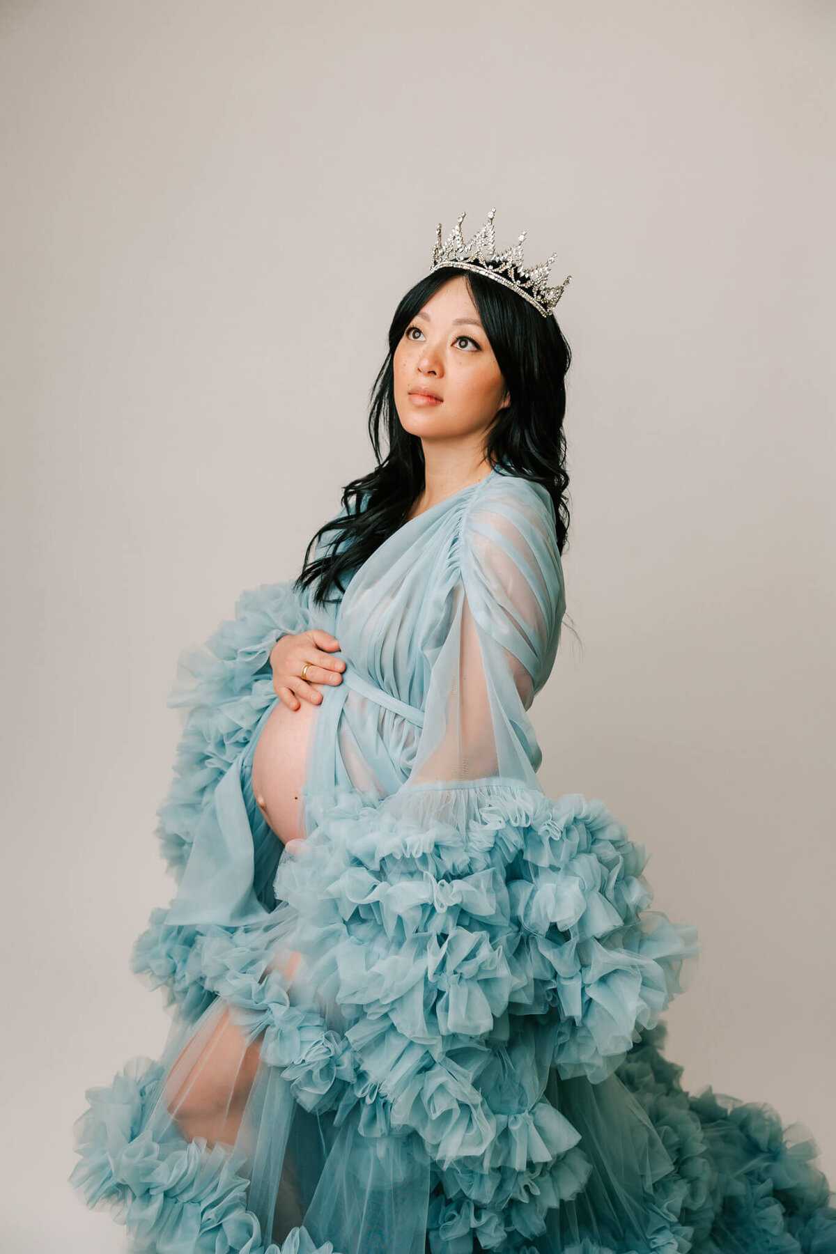 three quarter portrait of a mom wearing a blue tulle dress and crown. She is looking off to the side and the background is white