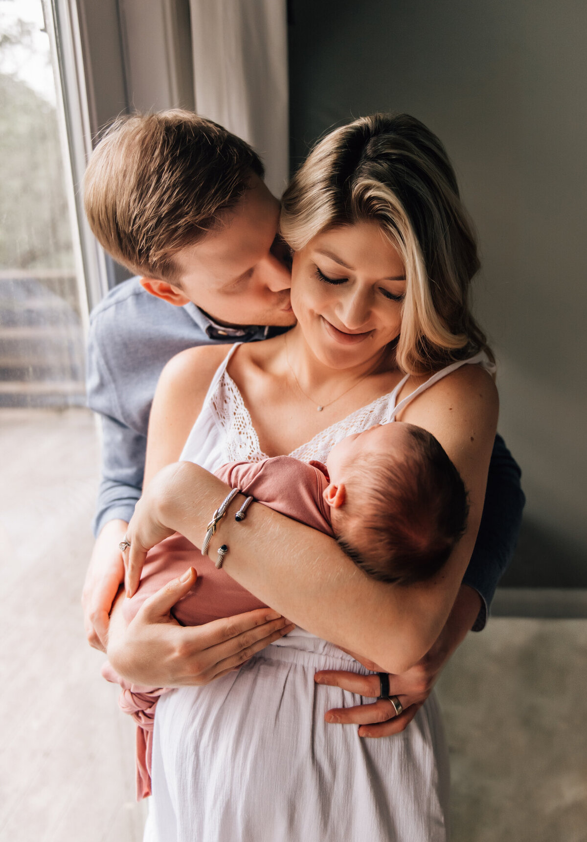 Newborn Photographer, Mom in dress holds baby daughter while dad kisses her on the side of her face.