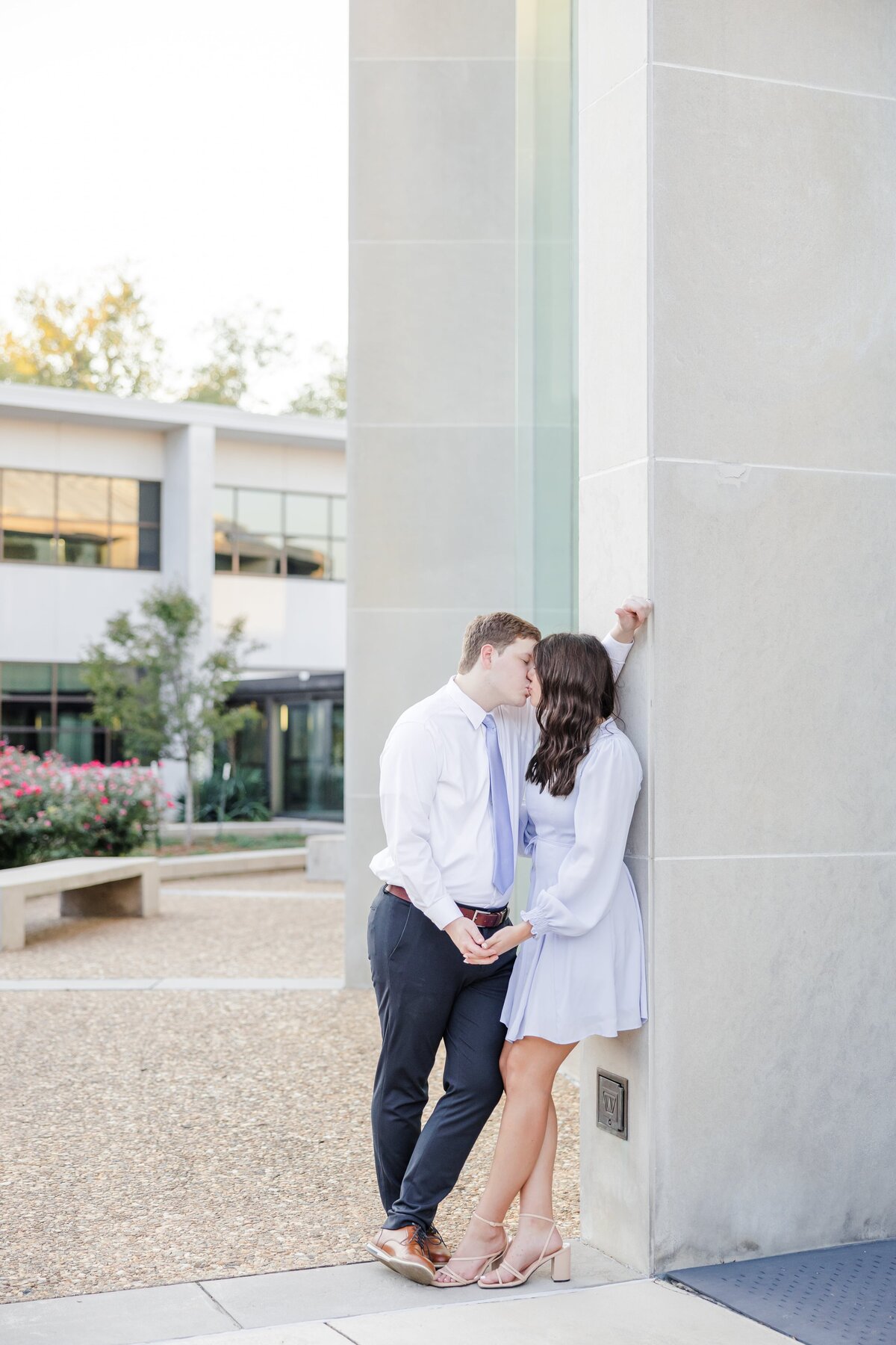 A women leans against a column in a blue dress kissing her fiancee in a matching tie