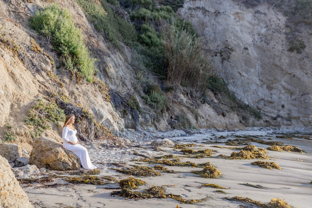 A woman in a white dress sits on a rock near a cliffside beach. The sandy shore is scattered with seaweed, and the cliffs are covered with green vegetation. The scene is serene and sunlit, with a natural coastal landscape.