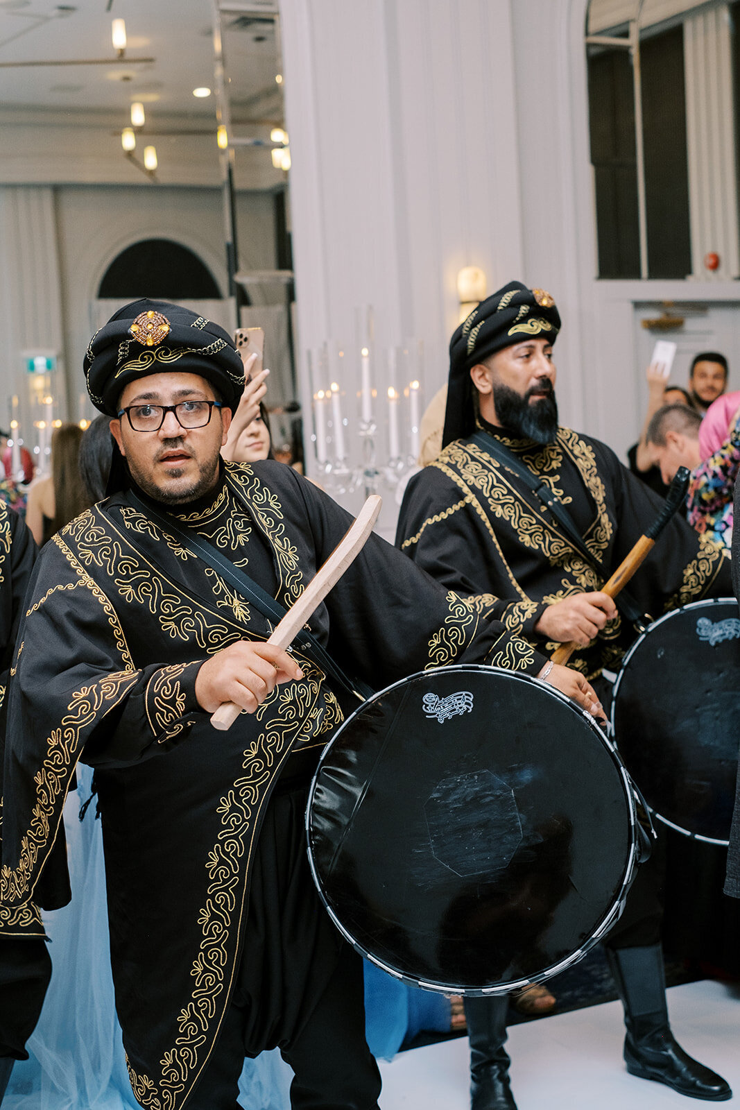 Two men in traditional attire play drums at an indoor event during a classic Calgary wedding. They wear black uniforms with gold embroidery and round hats. The background shows other attendees, candles, and decorative elements, capturing the essence of a cultural celebration at Fairmont Palliser Calgary.