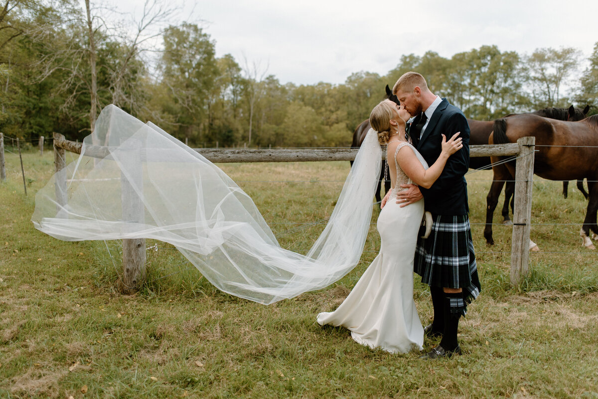 hamilton ontario wedding bride and groom kiss in front of horses behind fence with veil flowing in wind