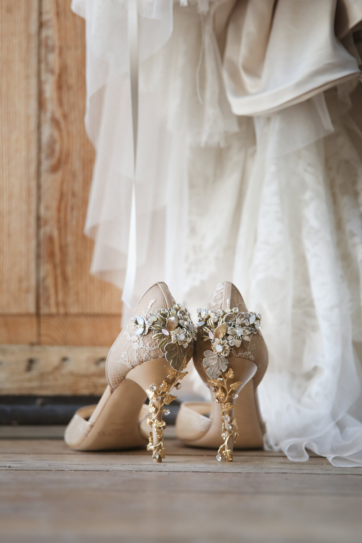 A pair of intricately designed bridal heels with gold floral accents sits beneath a flowing lace wedding gown, capturing timeless wedding elegance.