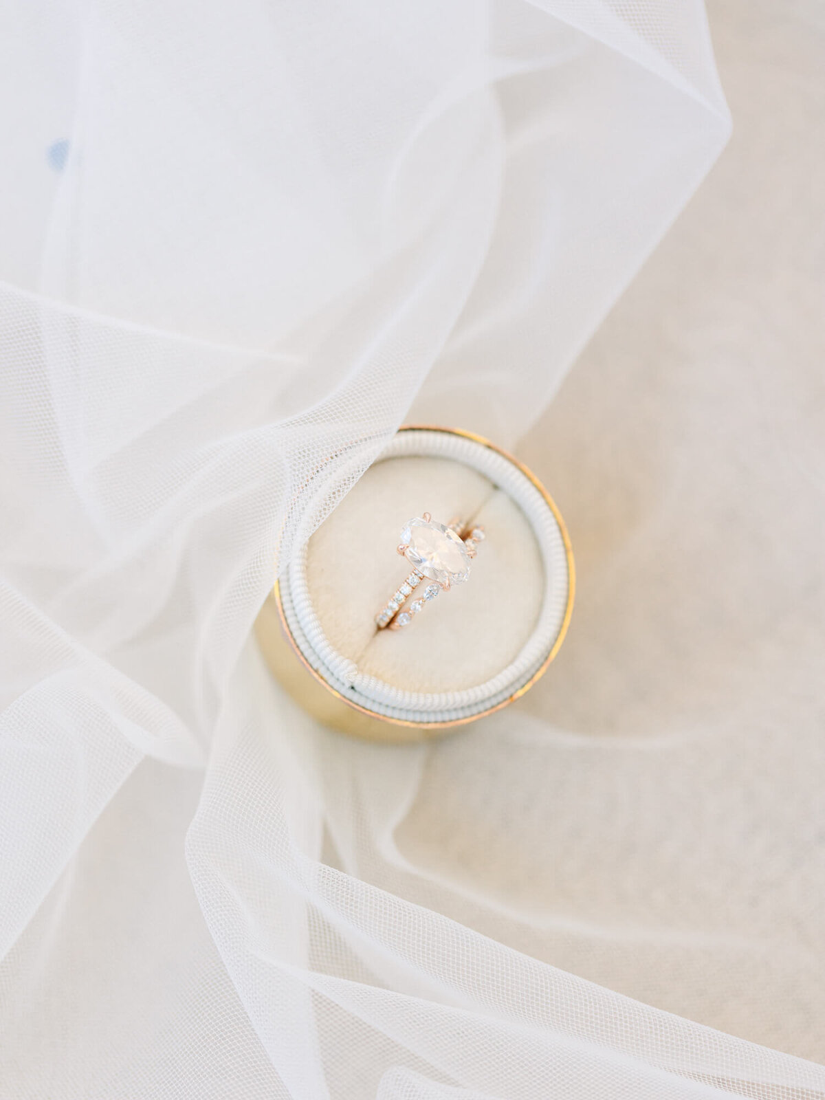 A delicate engagement ring with a large diamond rests in a cream-colored velvet box, partly draped by soft white tulle.