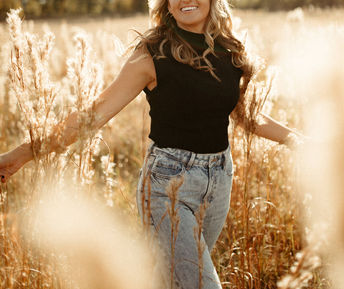 Senior girl with  curly hair wearing a black shirt and blue jeans frolicking through a field with tall grass