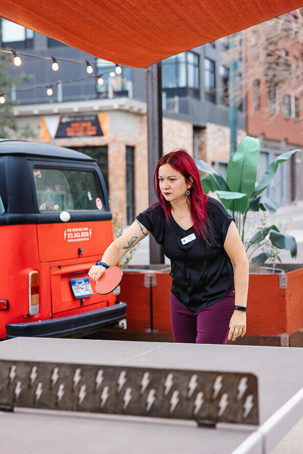 Woman plays ping pong with a decorative red bus behind her.