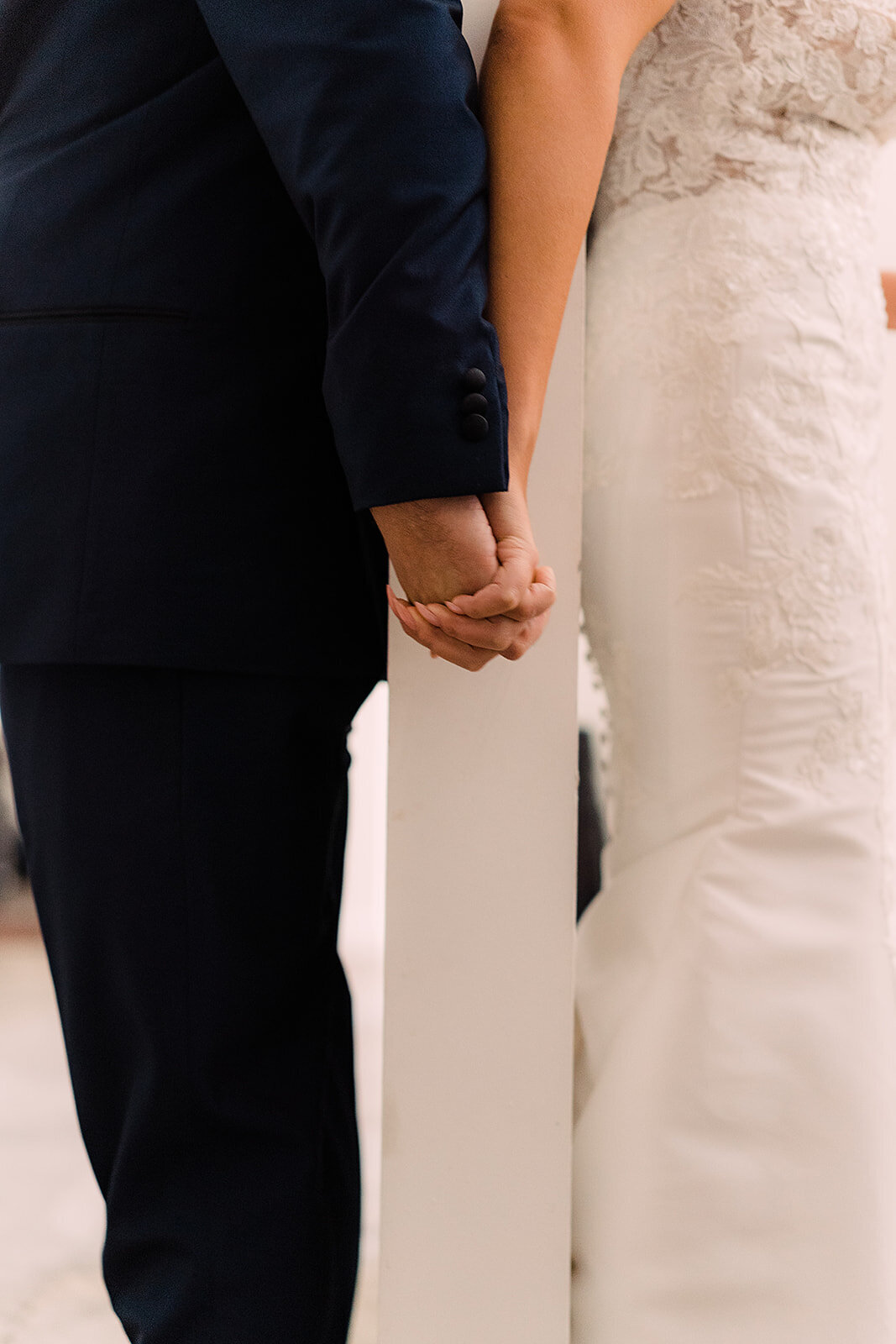Bride and groom hold hands separated by a wall, symbolizing the anticipation before seeing each other on their wedding day.