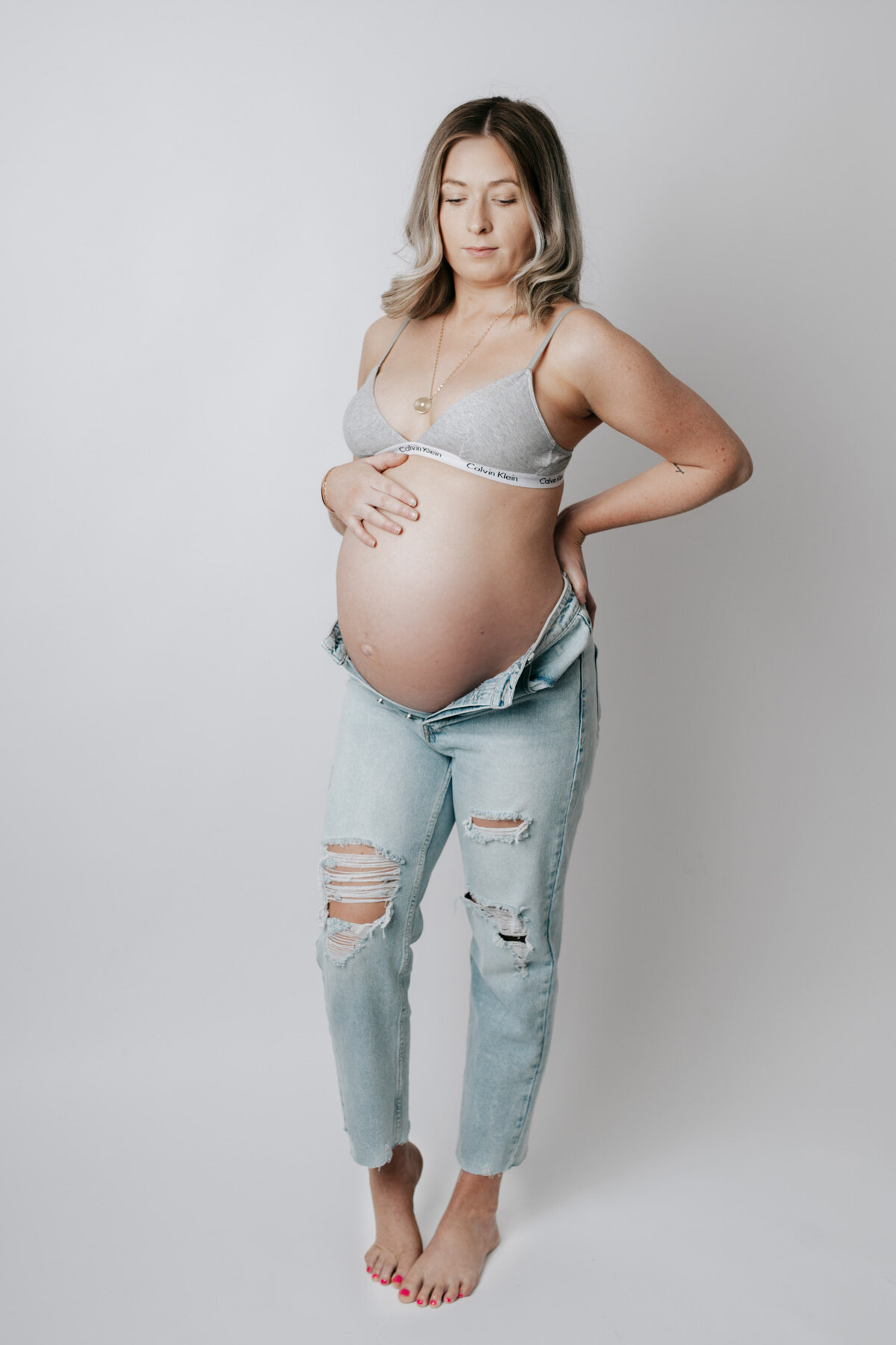 Studio Maternity session in Exeter, Ontario photography studio - mom in distressed ripped jeans unzipped exposing her baby bump. Mom is wearing a grey bralette and smiling at her bump against a white backdrop.