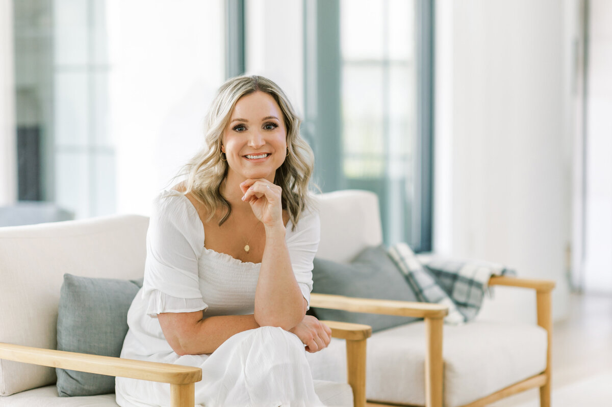 Top Ontario, Canada interior designer and decorator focused on stylish and functional designs poses for a headshot in her home. She is sitting on a cream upholstered chair with a wooden frame. One hand is resting under her chin and she is smiling at the camera.
