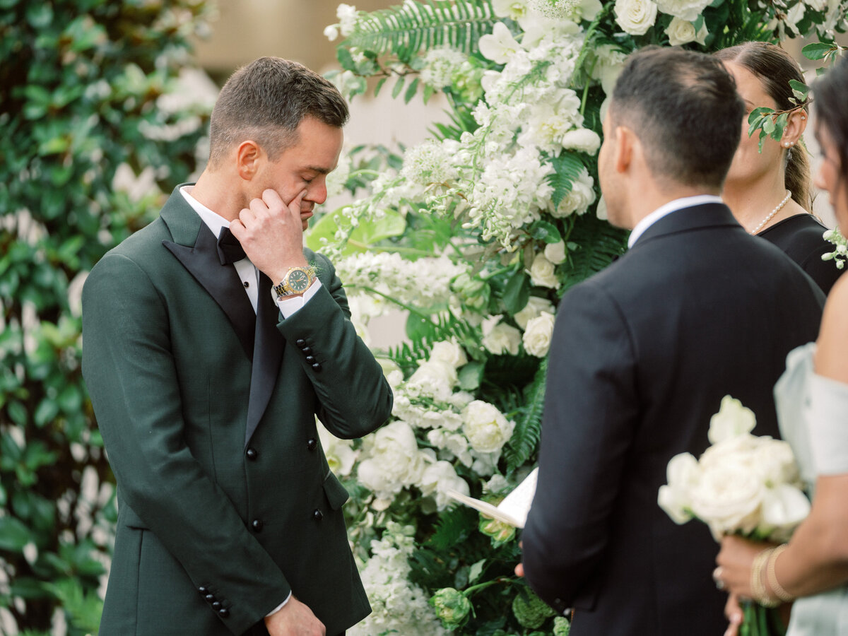 A groom displays deep emotion during a wedding ceremony surrounded by beautiful floral arrangements and an intimate gathering, capturing a heartfelt moment of love.