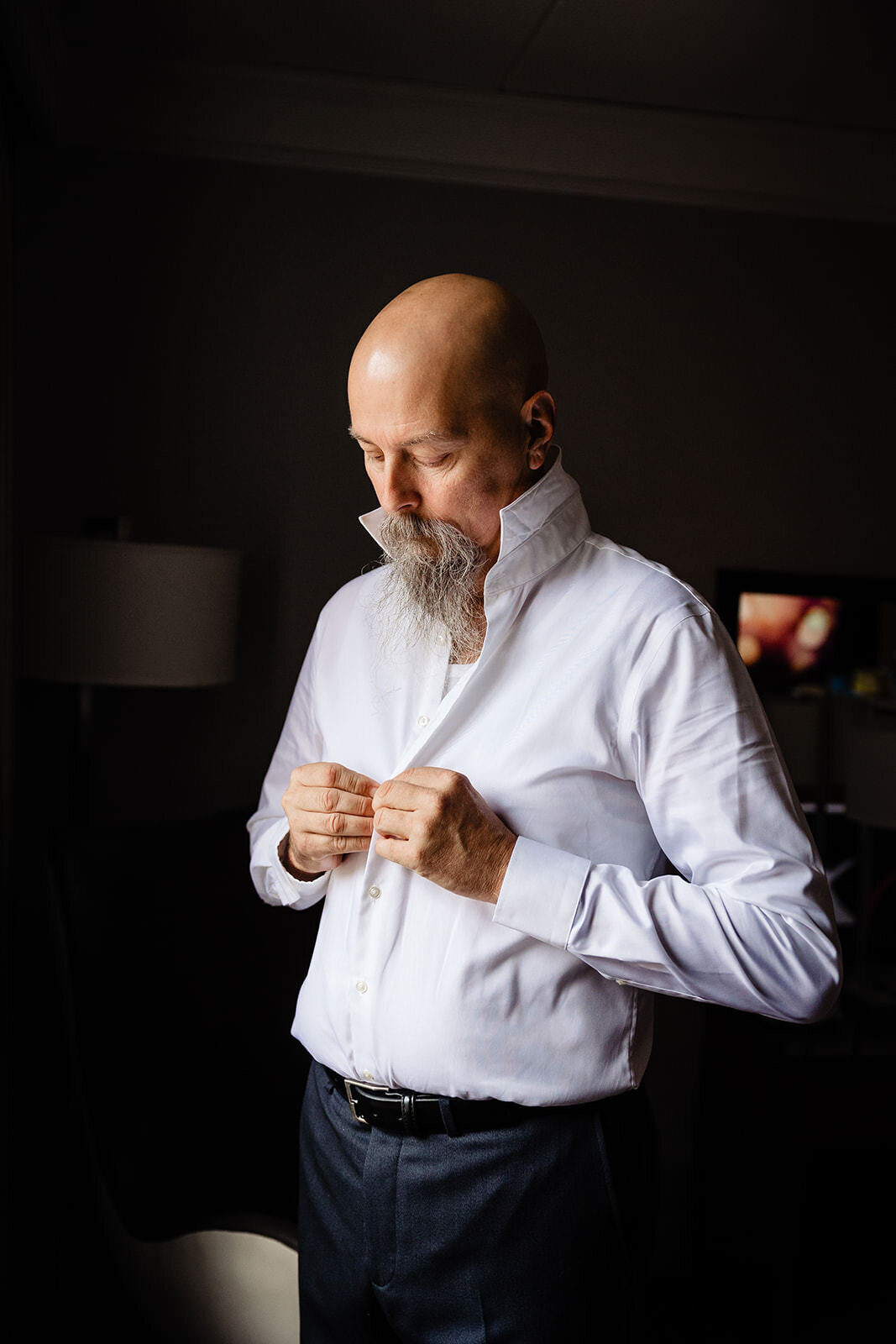 A man with a bald head and a full white beard buttoning up his white shirt, preparing for a formal event
