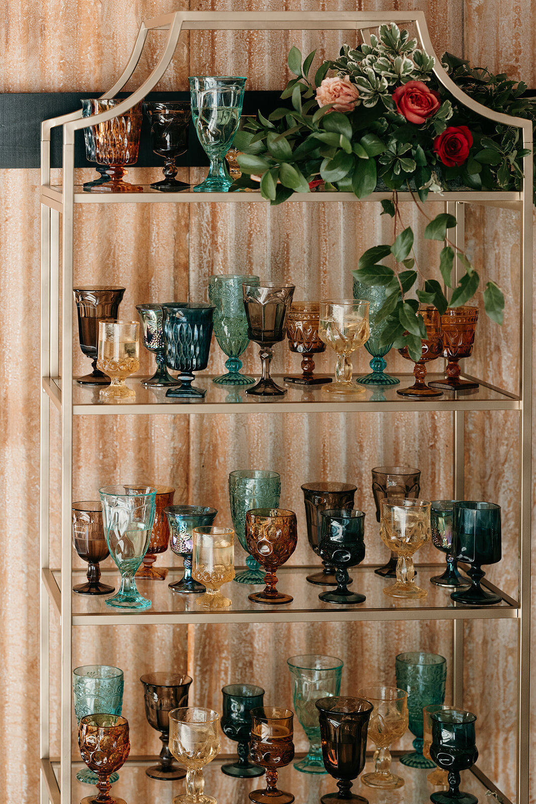 Shelf display of colorful vintage glassware in various shades and designs, adorned with greenery and flowers.