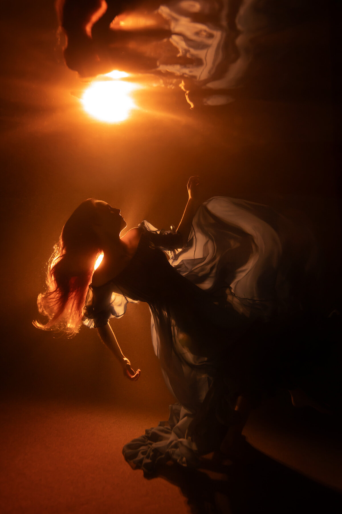 A woman in a flowing blue gown gracefully floats underwater, bathed in golden light that casts her silhouette against the darkened surroundings. Her hair glows with the warmth of the light, enhancing the ethereal mood of this Dallas underwater maternity photographer's captivating artwork.