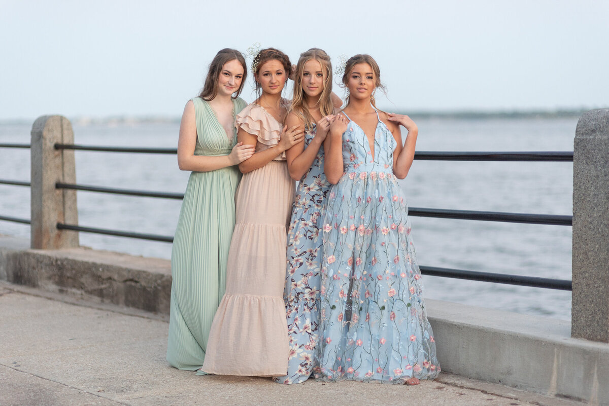 Four women wearing prom dresses, smiling next to the water